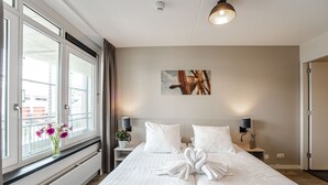 Habitación de hotel con una cama grande, una ventana con cortinas, una mesita de noche con lámpara y un cuadro colgado en la pared.