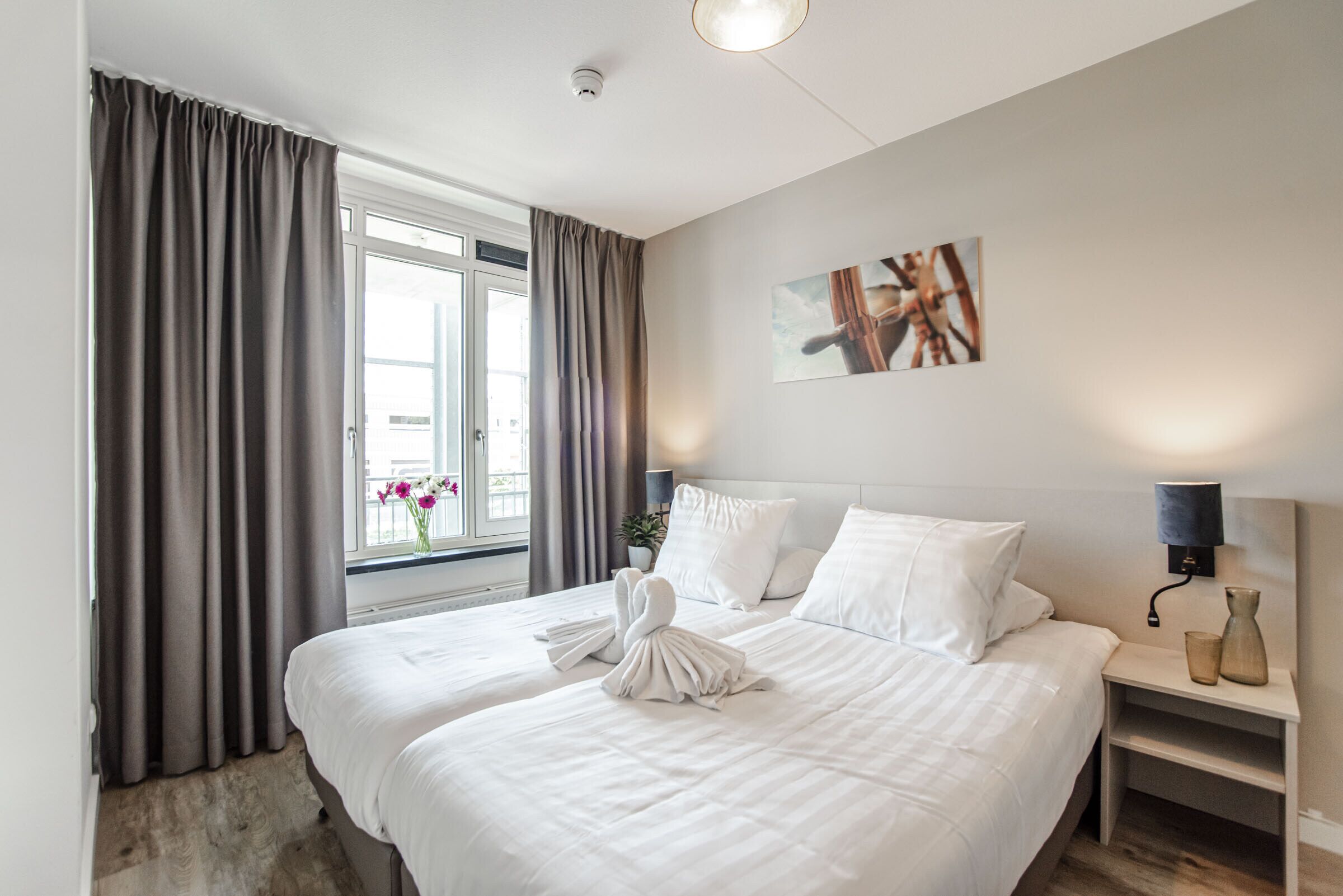 A hotel room with two beds, a large window with curtains, a wall-mounted artwork, and a bedside table with a lamp.