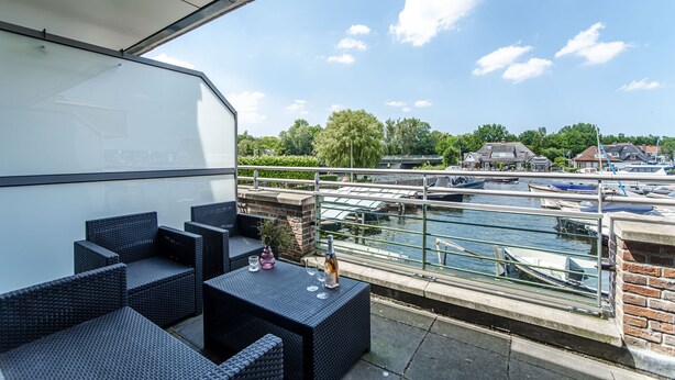 Un balcón con muebles de mimbre, una botella y vasos, con vistas a un canal con barcos y casas.