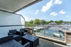 Un balcón con muebles de mimbre, una botella y vasos, con vistas a un canal con barcos y casas.