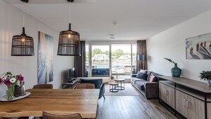 Una sala de estar moderna con una mesa de comedor de madera, lámparas colgantes negras, un sofá gris y un ventanal con vistas a edificios.