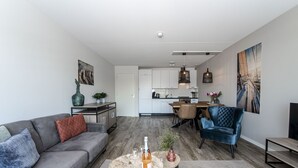 Una sala moderna con un sofá gris, una mesa de centro con una botella y vasos, un sillón azul y un comedor con una mesa y sillas de madera.