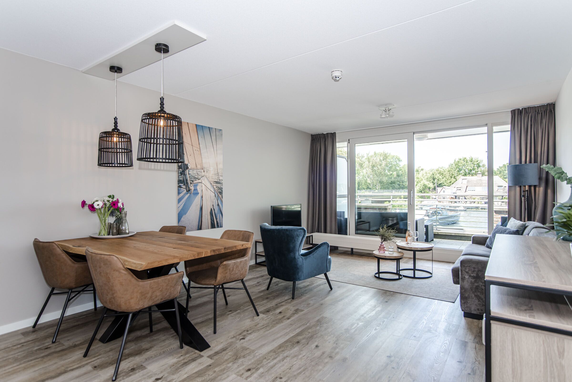 A modern living room with a wooden dining table, blue armchairs, a sofa, and a large window with a view of the city.