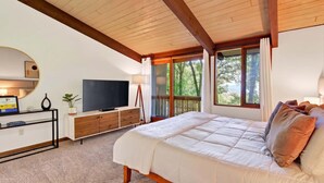 A bedroom with a bed, a TV, a desk with a computer, and a window with a view of trees.