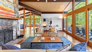 A spacious living area with a stone fireplace, wooden ceiling, and large windows offering a view of the outdoors.
