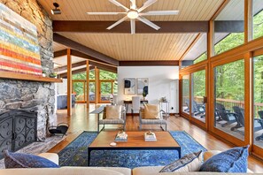 A spacious living area with a stone fireplace, wooden ceiling, and large windows offering a view of the outdoors.