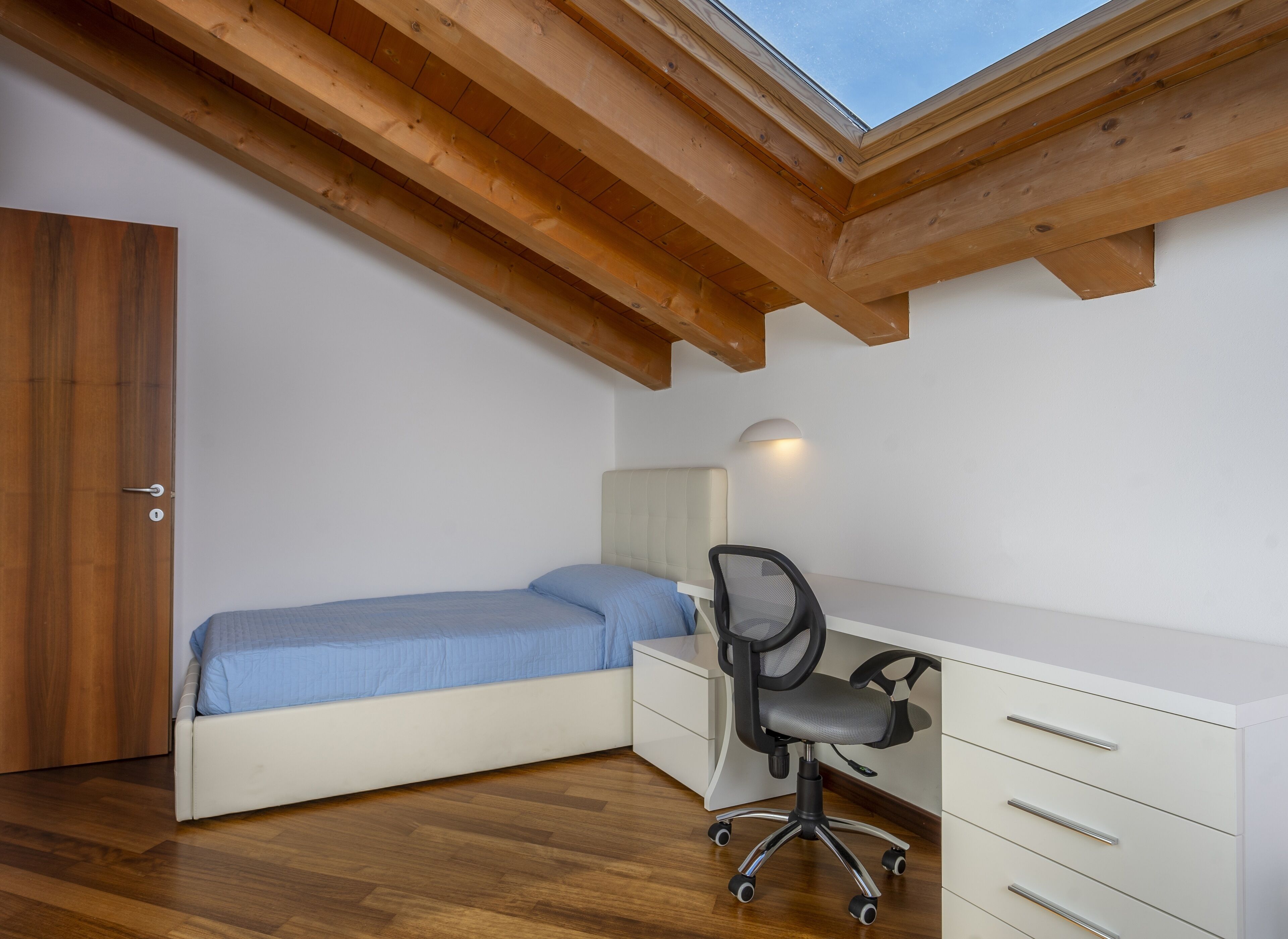 A bedroom with a bed, a desk, and a chair. There is a skylight above the bed.