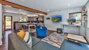Een moderne woonkamer met een bank, een salontafel en een televisie. Achtergrond toont een keuken met kastjes en een eethoek.