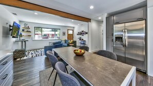 Een moderne keuken met een roestvrijstalen koelkast, een eethoek met een houten tafel en grijze stoelen, en een zithoek met een bank en een aan de muur bevestigde televisie.