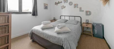 A neatly made bed with a grey patterned bedspread and pillows, a bedside table with a lamp, and a wall-mounted sign that reads "Explore World".