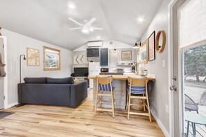 A modern kitchen with a central island, a dining area with two wooden chairs, a dark sofa, and a ceiling fan.