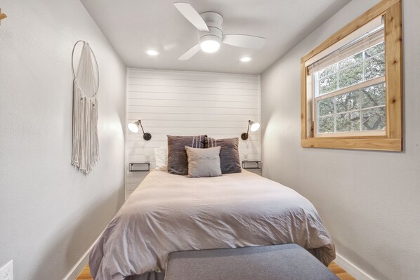 Un dormitorio con cama, lámparas de noche, ventilador de techo y ventana con vista a árboles.