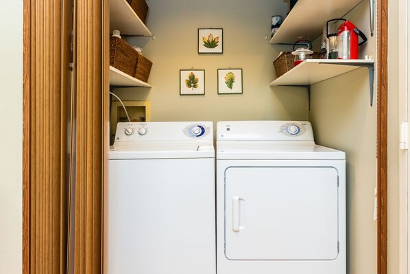 A laundry room with two white front-loading washers and dryers, wooden shelves with baskets, and three framed botanical prints on the wall.