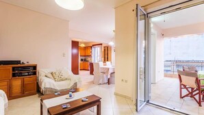 Sala de estar com sofá, mesa de centro e televisão. Porta de correr de vidro dá acesso a uma varanda com mobiliário de exterior.