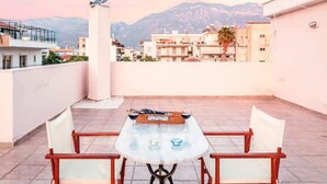 Terraço no telhado com mesa e cadeiras, uma escultura de pássaro e vista para edifícios e montanhas ao fundo.
