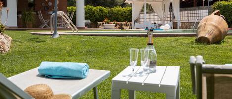 Zona junto a la piscina con sillones, una mesa con vasos y una botella, y una urna grande.