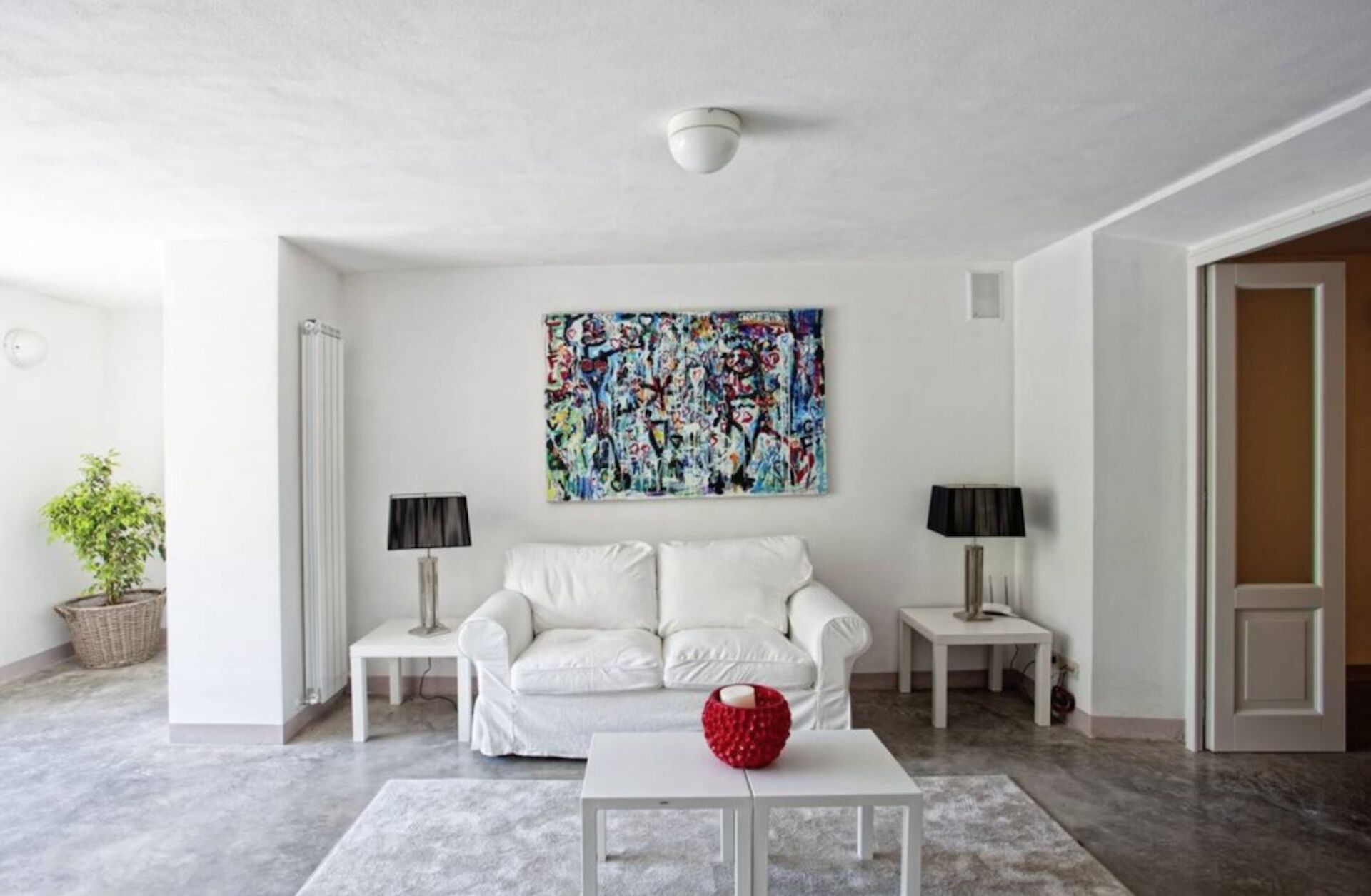 Ein modernes Wohnzimmer mit einem weißen Sofa, zwei Beistelltischen, einem Couchtisch und einem farbenfrohen, abstrakten Gemälde an der Wand.