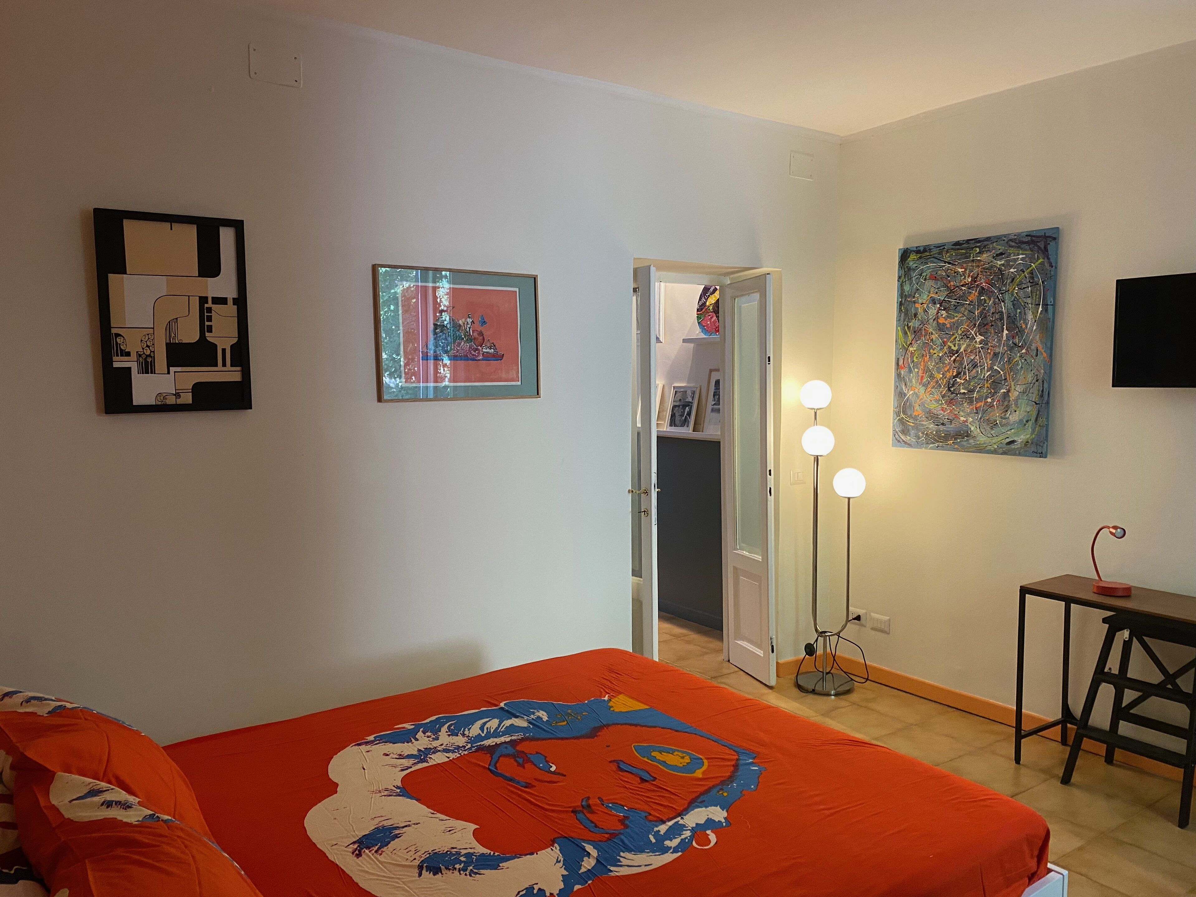 Ein Zimmer mit einem Bett, einem Schreibtisch und einer Tür zu einem weiteren Raum. An den Wänden hängen Bilder in Rahmen und eine Lampe mit drei Leuchten.