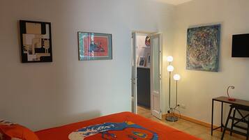 Ein Zimmer mit einem Bett, einem Schreibtisch und einer Tür zu einem weiteren Raum. An den Wänden hängen Bilder in Rahmen und eine Lampe mit drei Leuchten.