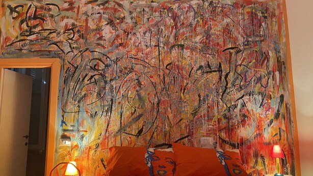 A bedroom with a large abstract painting on the wall, a bed with orange bedding, and a nightstand with a lamp.