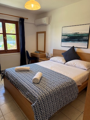 A hotel room with a bed, a desk, a chair, a window with curtains, an air conditioning unit, and a painting on the wall.