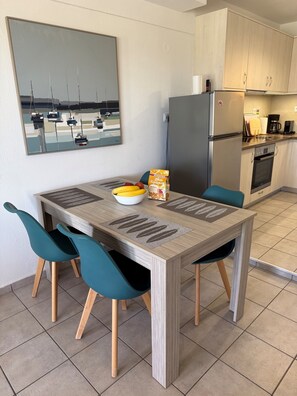 A modern kitchen with a refrigerator, stove, and coffee maker. There is a dining table with chairs and a bowl of fruit.