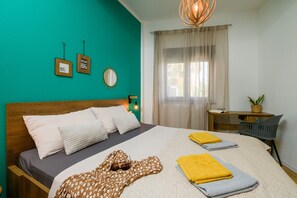 Une chambre avec un mur de couleur turquoise, une tête de lit en bois, un lit avec plusieurs oreillers, une table de chevet avec une lampe, une fenêtre avec des rideaux légers et un petit bureau agrémenté d’une plante.