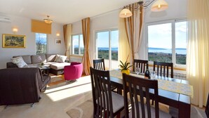A modern living room with a sofa, a dining table with chairs, and a view of the sea through large windows.