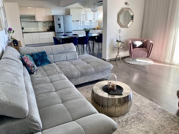 Un salon moderne avec un canapé d’angle gris, une table basse ronde et un fauteuil rose. On aperçoit une cuisine à l’arrière-plan.