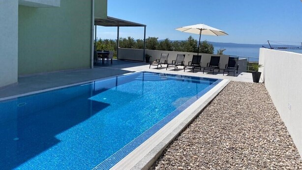 A rectangular swimming pool with a clear blue water surface, surrounded by a pebble-lined deck. There are lounge chairs and umbrellas arranged for relaxation. - Villa Noura in Makarska, private pool,gym (Makarska)