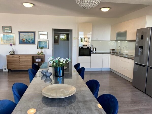 A modern kitchen with a dining area, featuring a long table, blue chairs, a refrigerator, and a microwave.