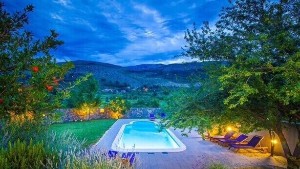 A swimming pool with lounge chairs and a stone pathway leading to it, surrounded by greenery and hills in the background.