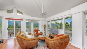 A spacious living area with wicker furniture, a ceiling fan, and large windows offering views of greenery. - Villas Of Kamalii 17 3 Bedroom Condo by RedAwning (Princeville)