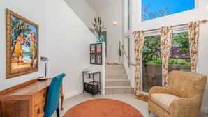 A spacious living area with a staircase leading to a balcony, a wicker chair, a desk with a chair, and a painting on the wall. - Villas Of Kamalii 17 3 Bedroom Condo by RedAwning (Princeville)
