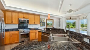 A modern kitchen with granite countertops, wooden cabinets, stainless steel appliances, and a large window offering a view of greenery. - Villas Of Kamalii 17 3 Bedroom Condo by RedAwning (Princeville)