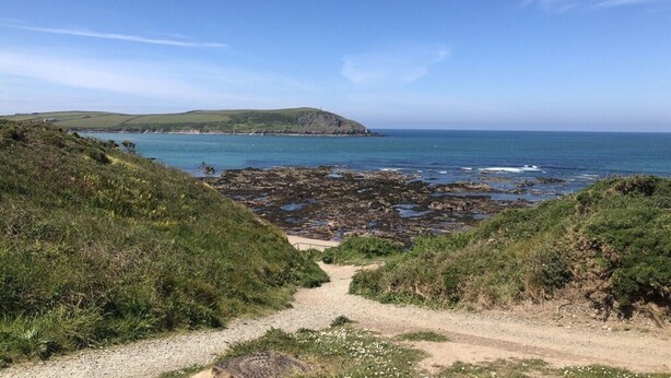 A coastal path leading to rocky shores with a clear view of the sea and distant hills.