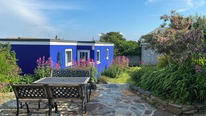 A stone-paved patio with a table and chairs, a blue building with windows, and surrounding greenery.