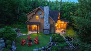 A log cabin with a stone chimney, surrounded by a well-maintained garden with red chairs and a stone pathway. - Waterfront - Family, Pool & Spa (Mille-Isles)