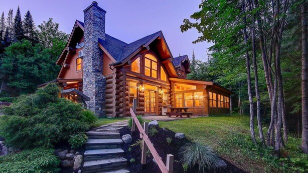 A log cabin with a stone chimney, large windows, and a stone staircase leading to the entrance. - Waterfront - Family, Pool & Spa (Mille-Isles)