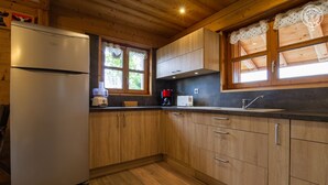 A kitchen with wooden cabinets, a refrigerator, a coffee maker, and a window with lace curtains.