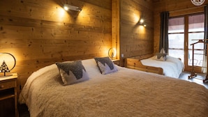 A cozy wooden room with two beds, each with a fluffy white bedspread and decorative pillows. There are wall-mounted lights and a window with curtains.