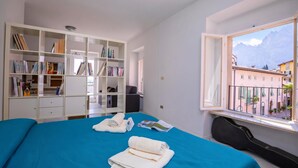 Ein Schlafzimmer mit Bett, Bücherregal, Fenster und Blick auf Gebäude, sowie einer Gitarre.