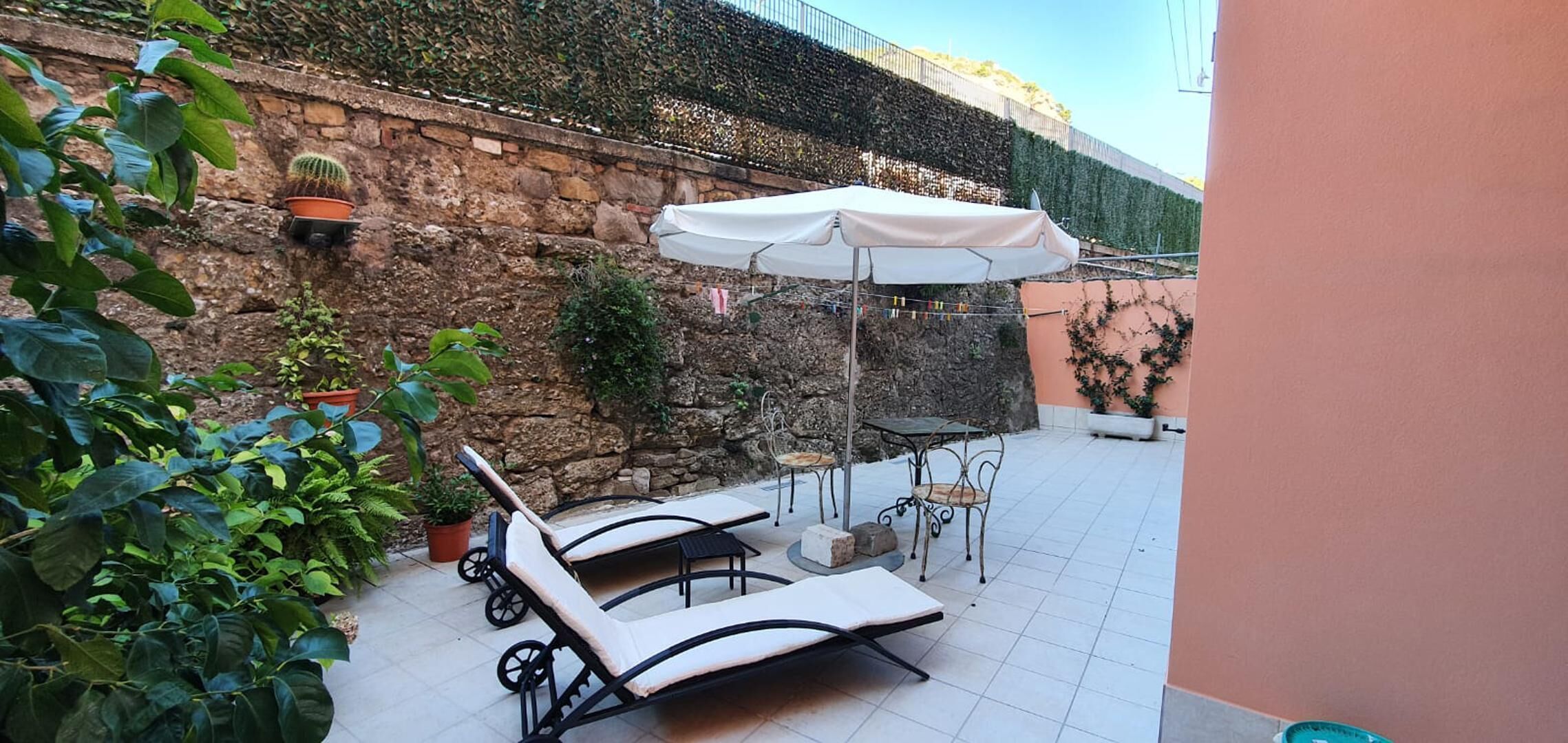 Une terrasse avec des fauteuils et une table, entourée de plantes en pot et d’un mur de pierre.