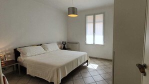 A bedroom with a bed, bedside table, lamp, and a window with blinds. - 1 Bed Flat - Sleeps 2 - Parking - Wifi (San Biagio della Cima)