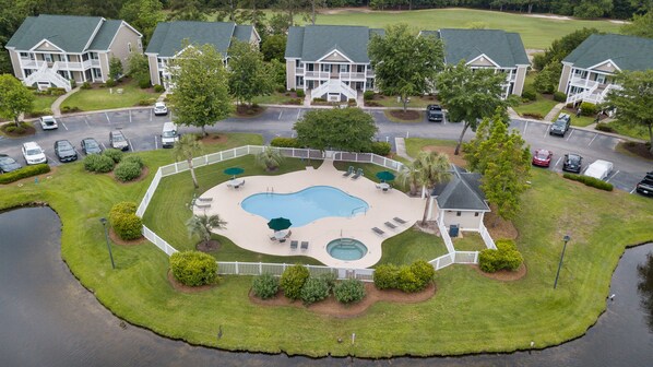 A residential area with a swimming pool surrounded by a fence, multiple houses, and parked cars.