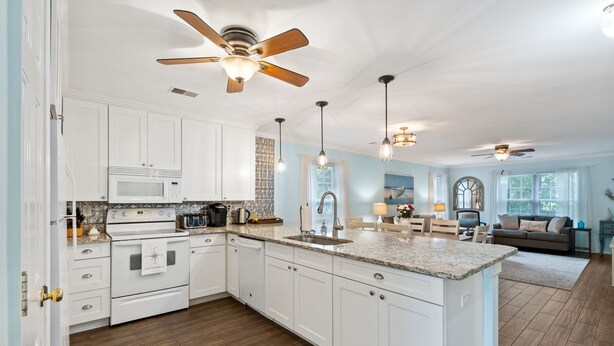 A spacious kitchen with white cabinets, a granite countertop, and a central island. There are ceiling fans, pendant lights, and a large window providing natural light. - Salt & Light - 1st Floor 3BR unit in True Blue (Pawleys Island)
