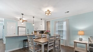 A modern kitchen with a central island, white cabinetry, and a dining area with a wooden table and chairs. - Salt & Light - 1st Floor 3BR unit in True Blue (Pawleys Island)