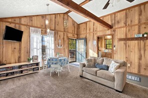 A cozy living room with a sofa, a table with chairs, a bookshelf, and a television mounted on the wall.