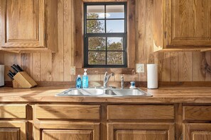 A kitchen with wooden cabinetry, a double sink, and a window with a view of trees.