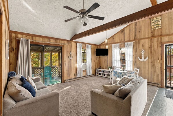 A spacious living room with a ceiling fan, wooden walls, a large window with curtains, a television mounted on the wall, and a cozy seating area with a sofa and chairs.